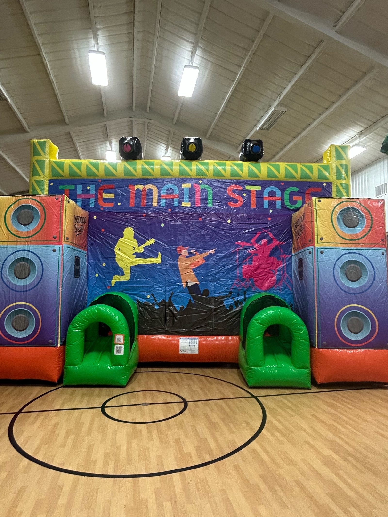 The Main Stage concert-themed bounce house with speaker towers and green arch entrances on a gym basketball court in Howe, TX